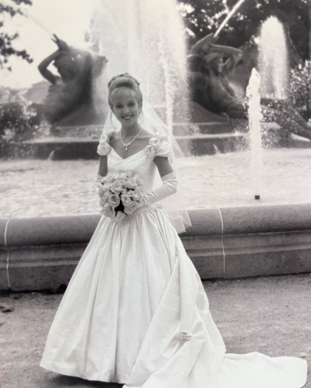 Bride in a full white wedding dress holding roses before a water fountain