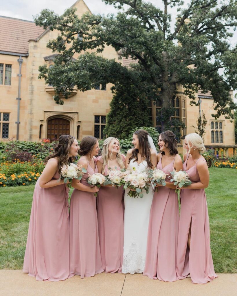 Bride and bridesmaids in dusty rose dresses holding bouquets before a stone manor.