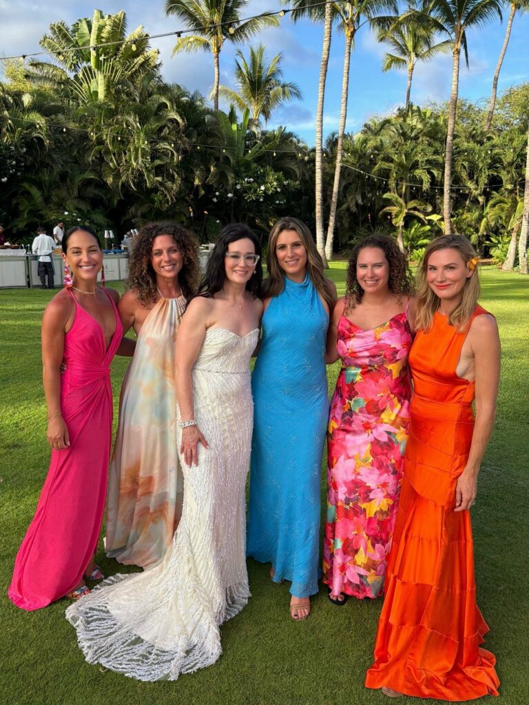 Six women in colorful formal gowns posing on a lawn with palm trees.