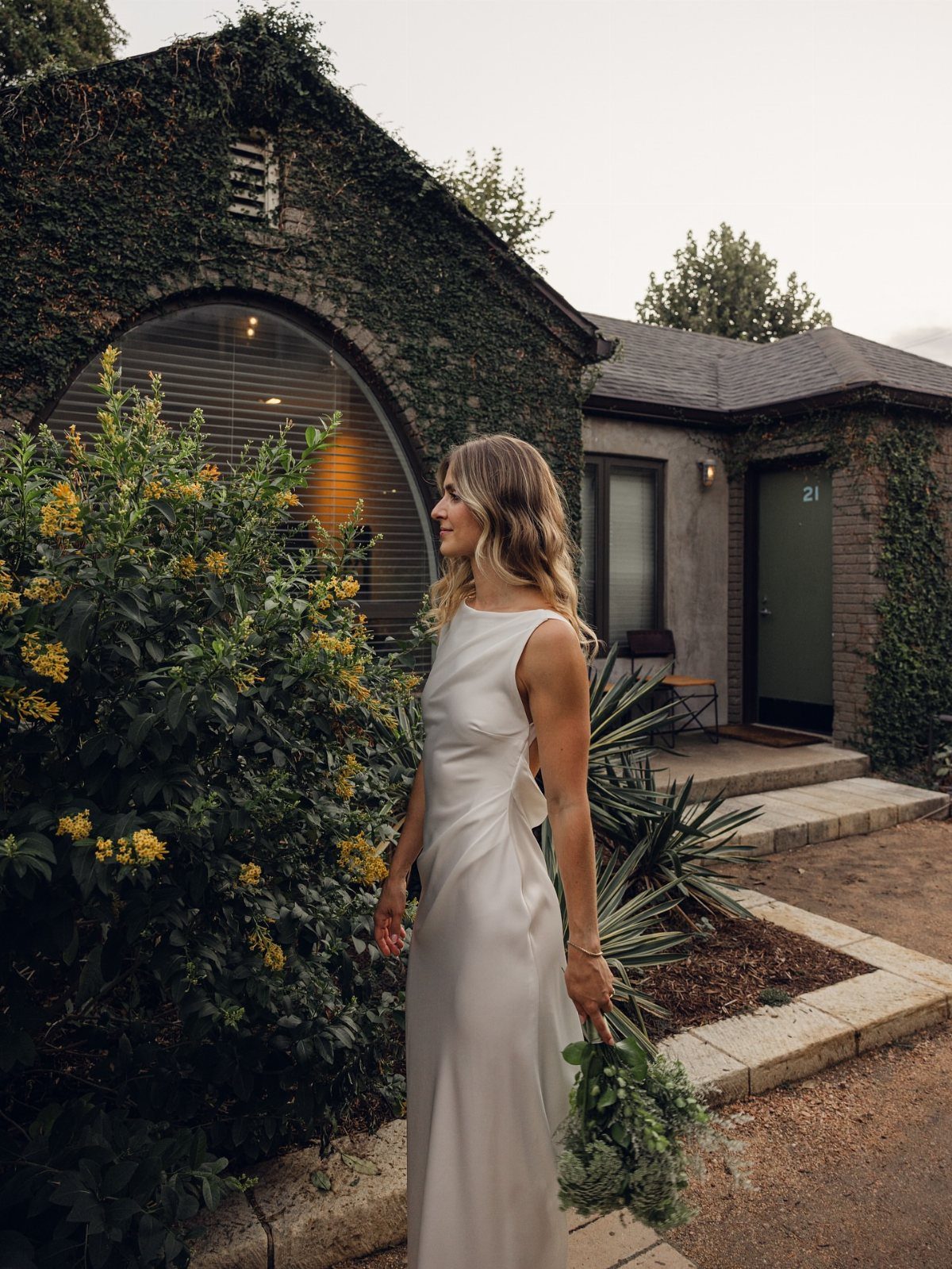 Woman in a sleek white minimalist wedding gown holding a green bouquet outdoors.
