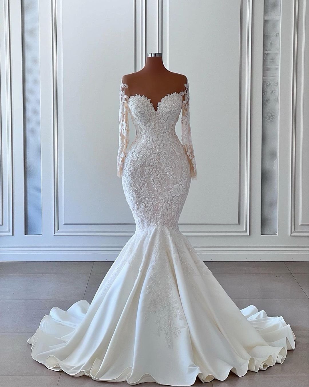 White lace mermaid bridal gown with long sleeves on a mannequin against white walls.
