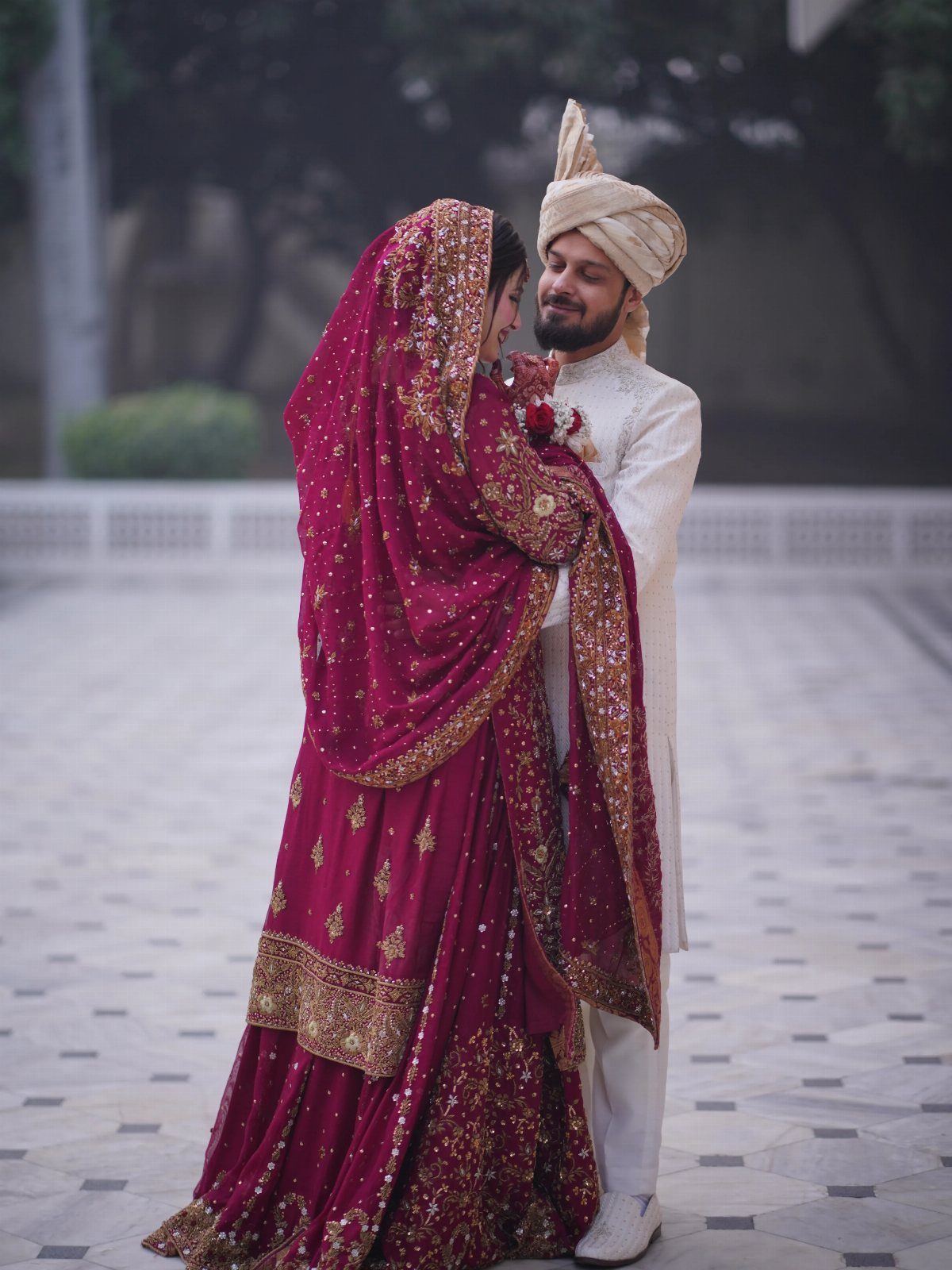 Bride in maroon lehenga and groom in white sherwani embracing in a courtyard