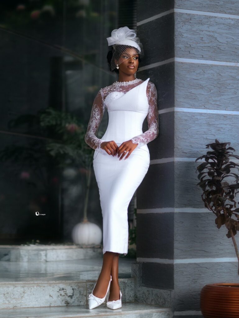 Woman in a white lace-sleeved midi wedding dress and fascinator posing by a wall