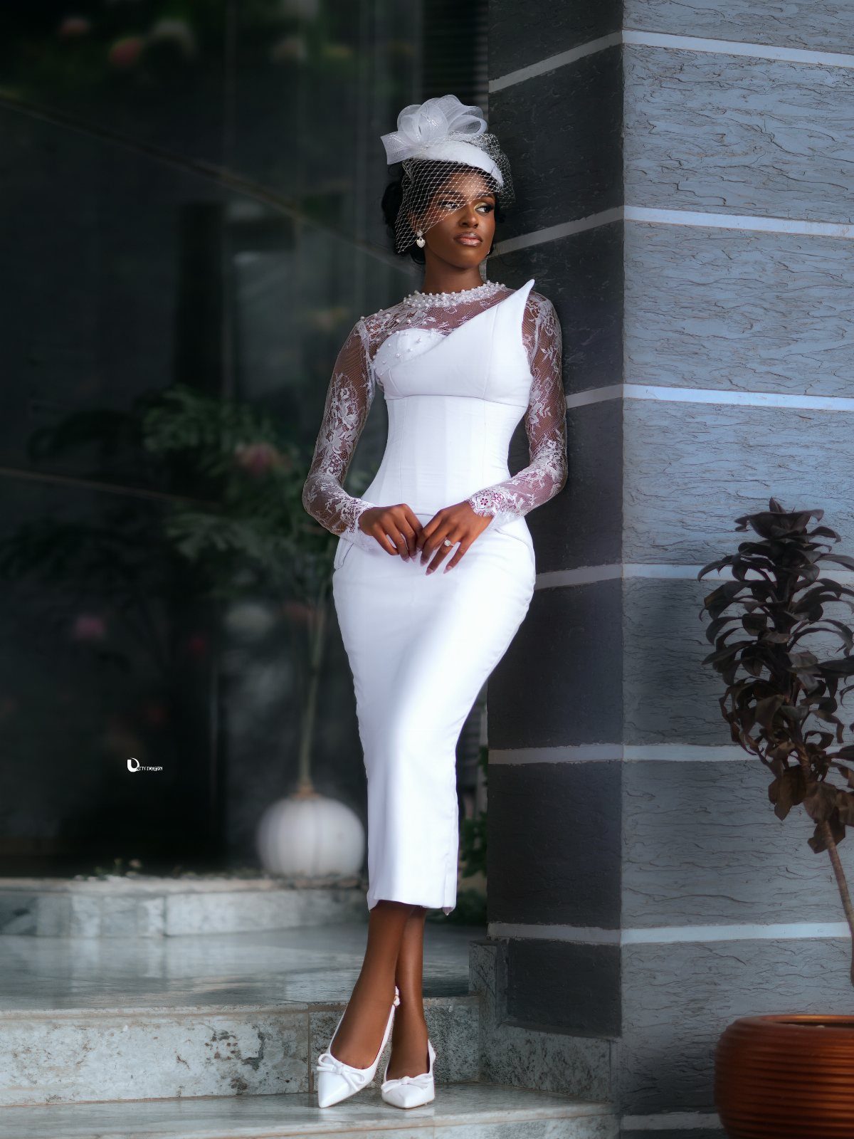 Woman in a white lace-sleeved midi wedding dress and fascinator posing by a wall