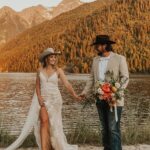 Bride in lace wedding dress and cowboy hat holding hands with groom by lake.