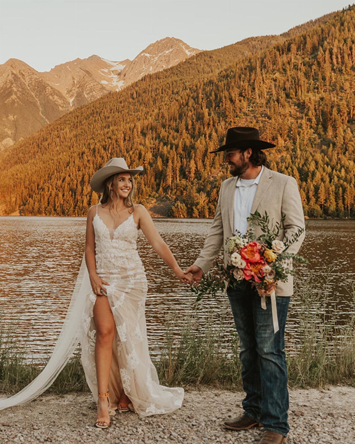 Bride in lace wedding dress and cowboy hat holding hands with groom by lake.