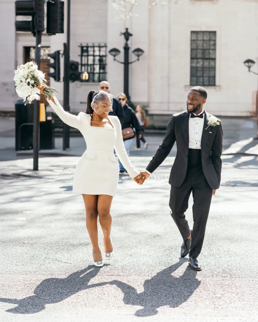 Couple in wedding attire walking hand-in-hand across a city street with white bouquet