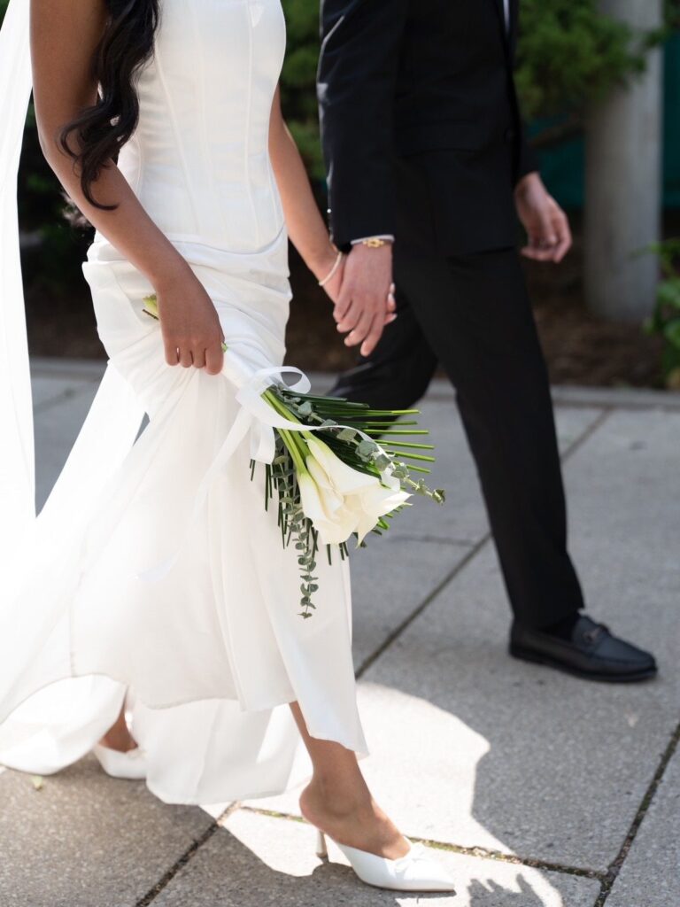 Woman in a white wedding dress holding a calla lily bouquet and partner's hand