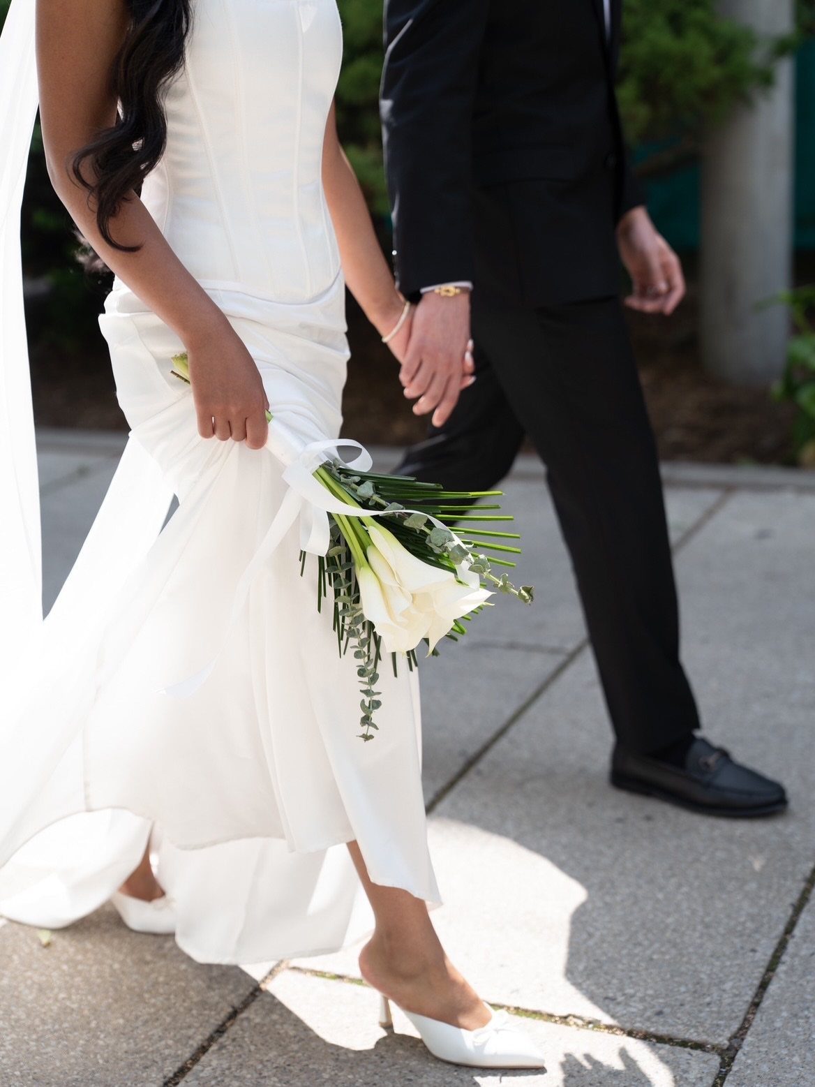 Woman in a white wedding dress holding a calla lily bouquet and partner's hand