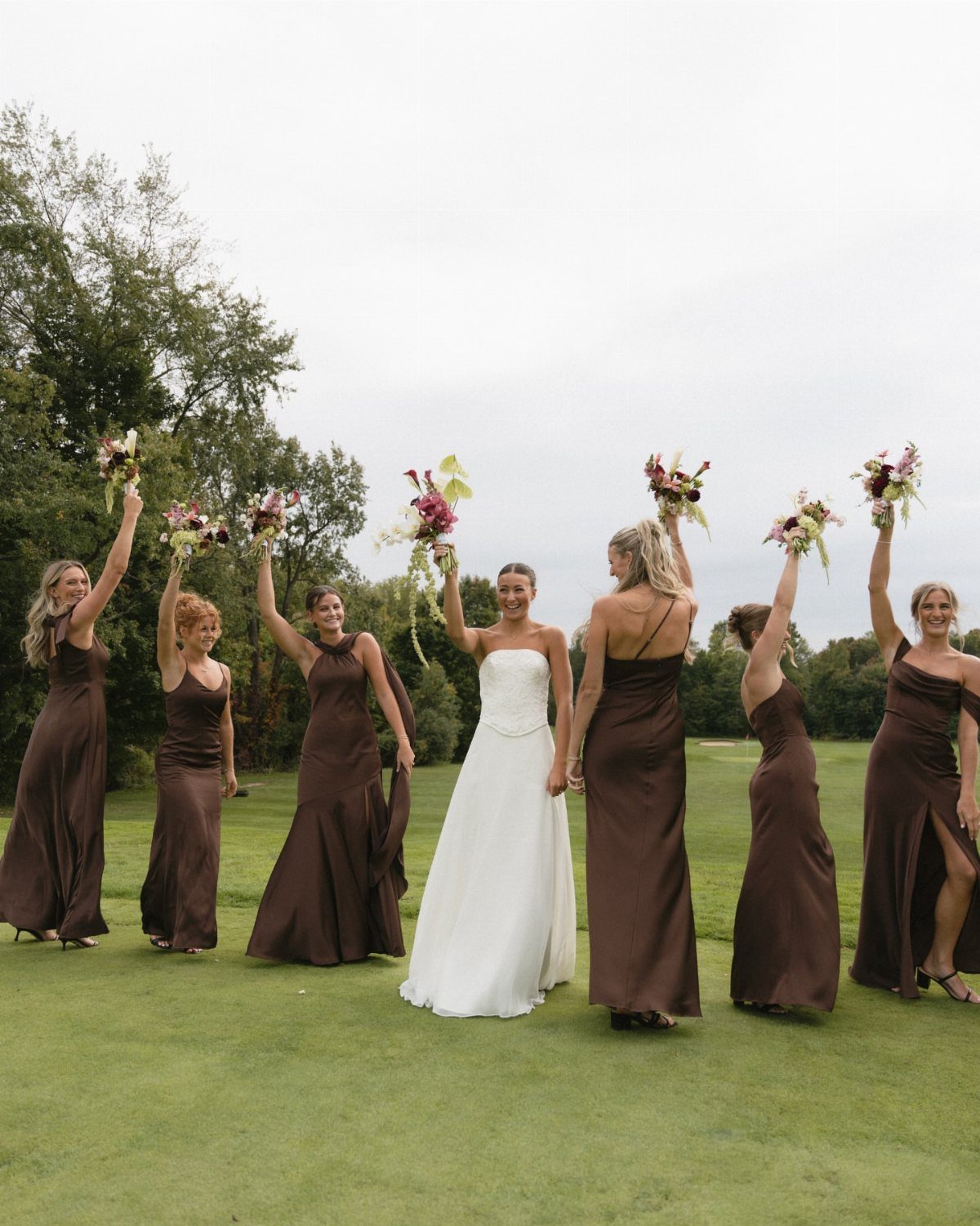Bride in white gown with bridesmaids in brown dresses holding bouquets on grass