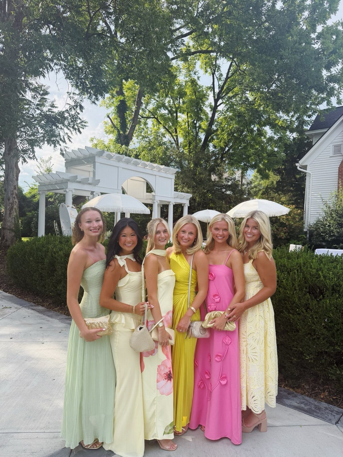 Six women in colorful formal gowns posing in a garden with a white pergola.