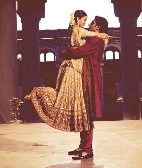 Groom in maroon sherwani lifting bride wearing a gold embroidered lehenga outdoors