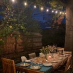 Outdoor dinner table set with plates and flowers under glowing string lights at dusk