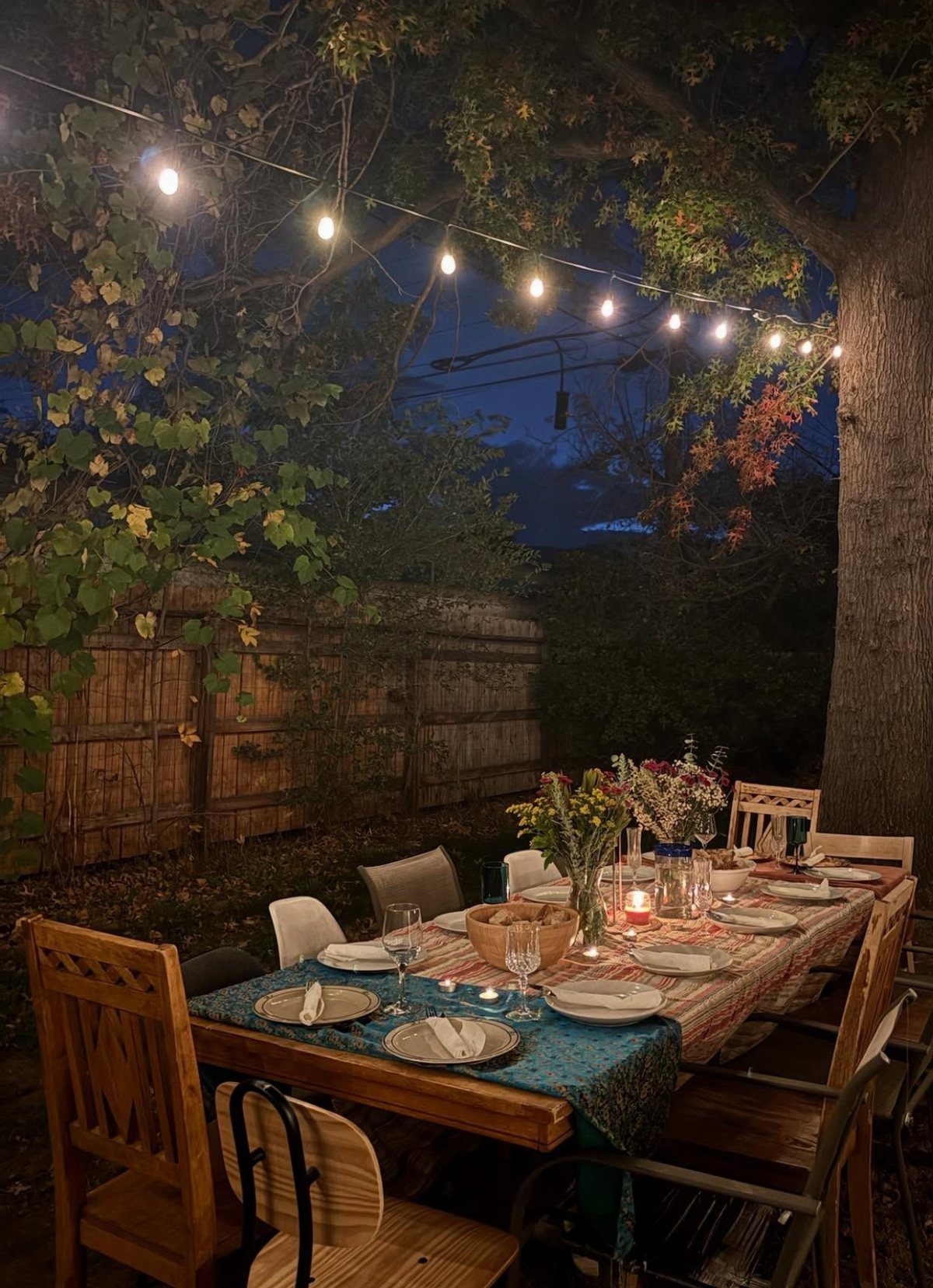 Outdoor dinner table set with plates and flowers under glowing string lights at dusk
