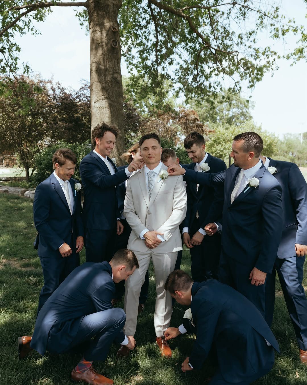 Groom in a white suit surrounded by groomsmen in navy blue suits outdoors.