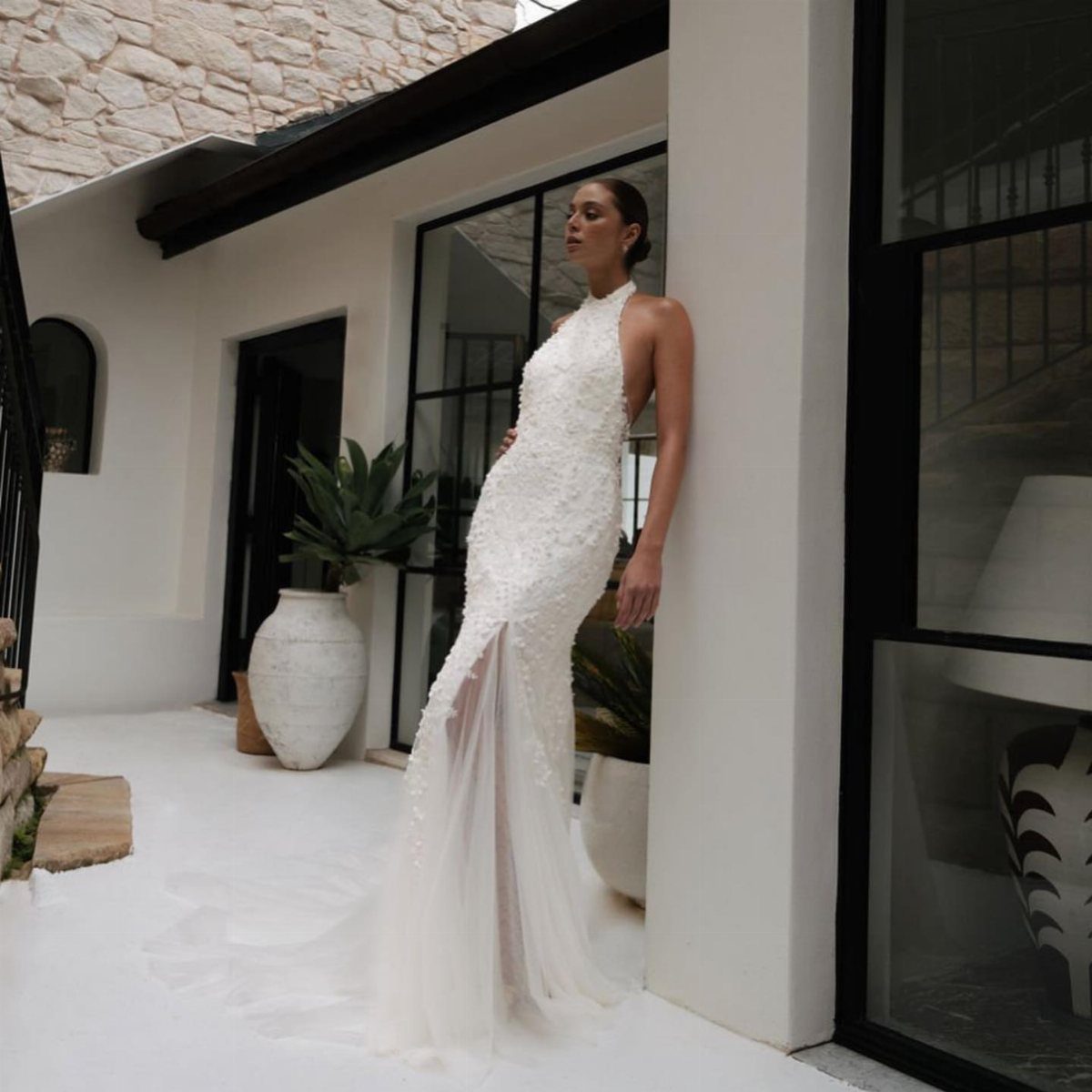 Woman in a white lace halter wedding dress leaning against a white wall