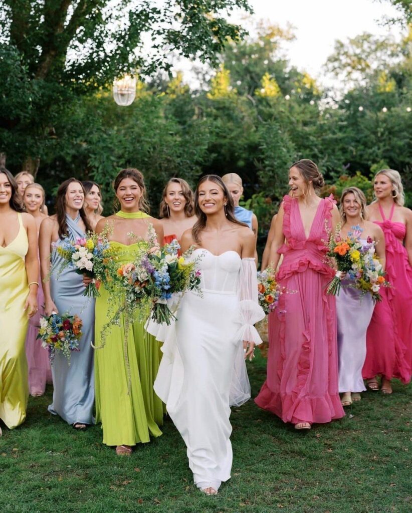 Bride in white gown walking with bridesmaids wearing mismatched colorful long dresses.