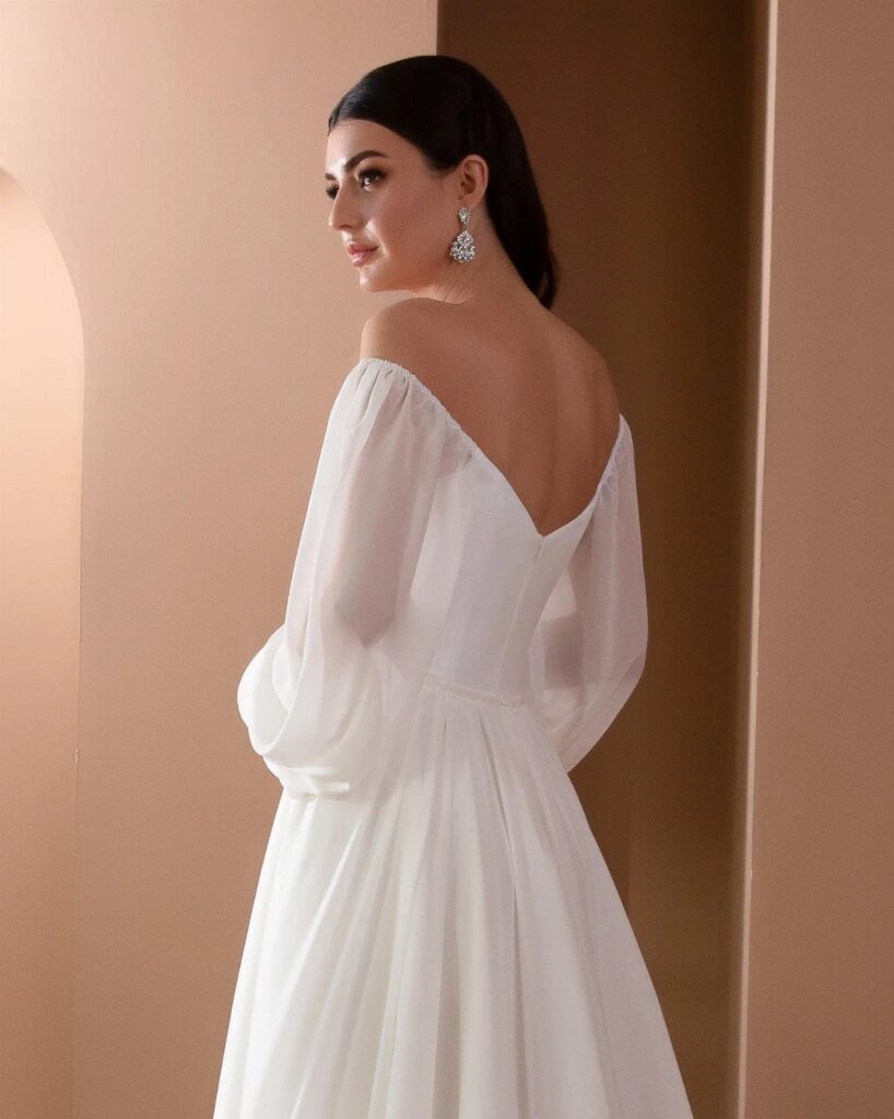 Woman in a white off-the-shoulder bridal dress with sheer sleeves against beige walls