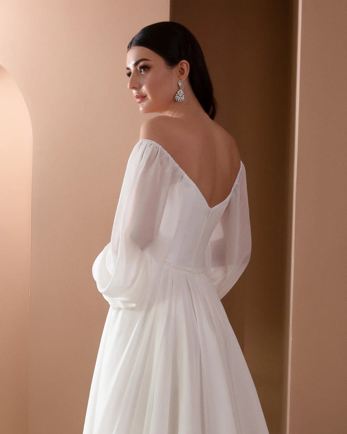Woman in a white off-the-shoulder bridal dress with sheer sleeves against beige walls