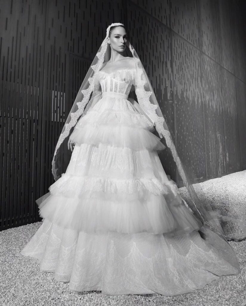 Woman in a tiered ruffle wedding dress and long lace veil standing outdoors.