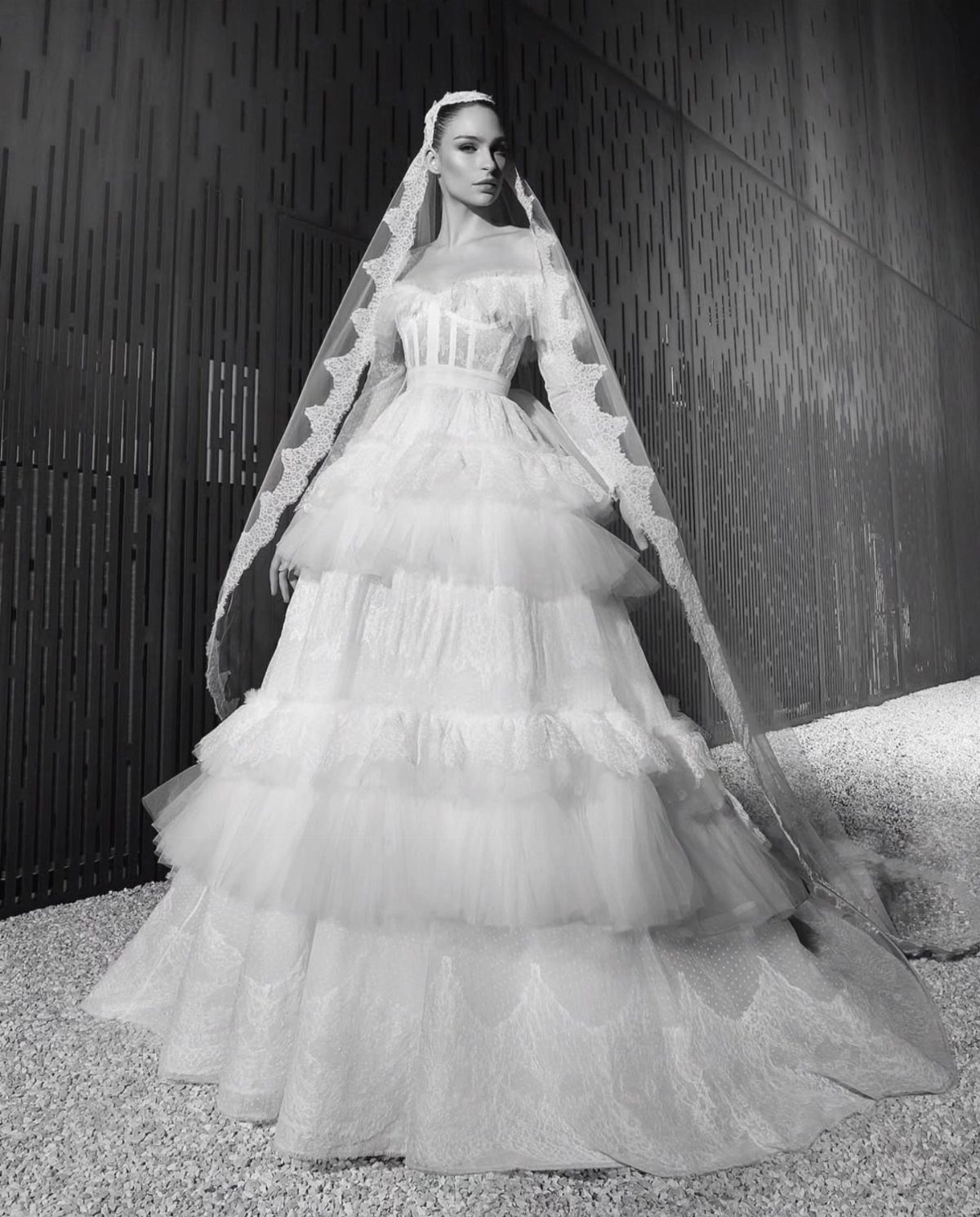 Woman in a tiered ruffle wedding dress and long lace veil standing outdoors.