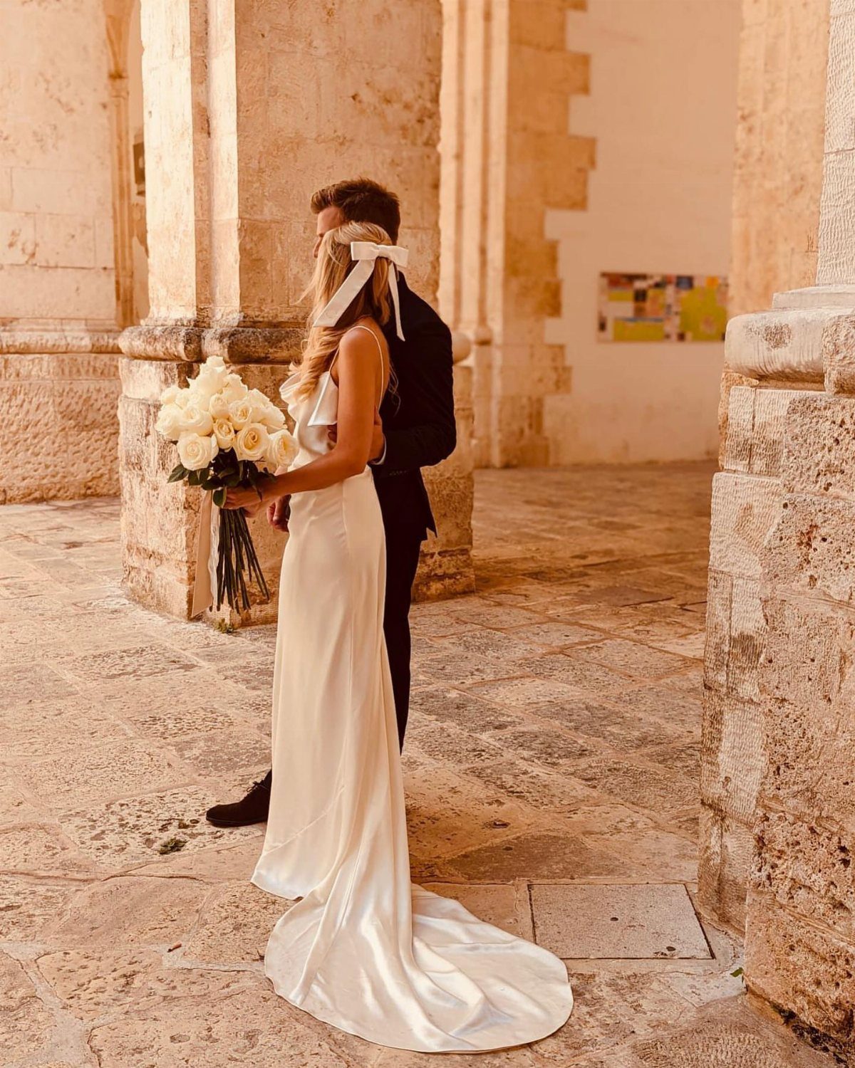 Woman in a white satin slip wedding dress holding white roses beside a groom.