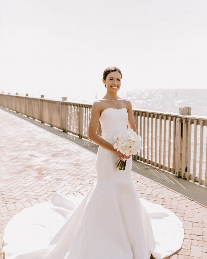 Woman in a white strapless wedding dress holding a bouquet on a beach boardwalk