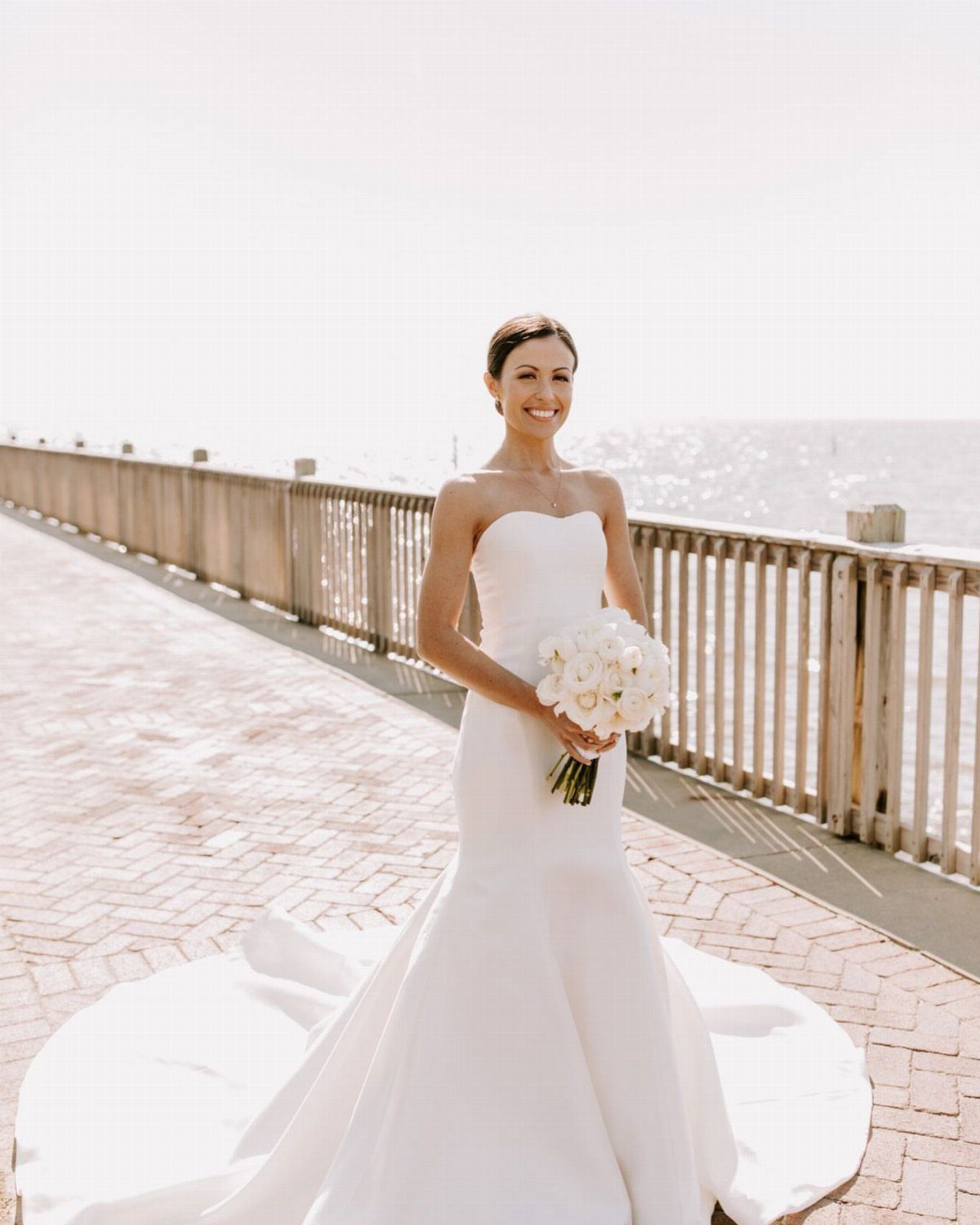 Woman in a white strapless wedding dress holding a bouquet on a beach boardwalk