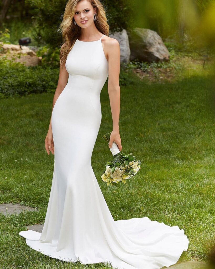Woman in a sleek white form-fitting wedding dress holding a white flower bouquet.