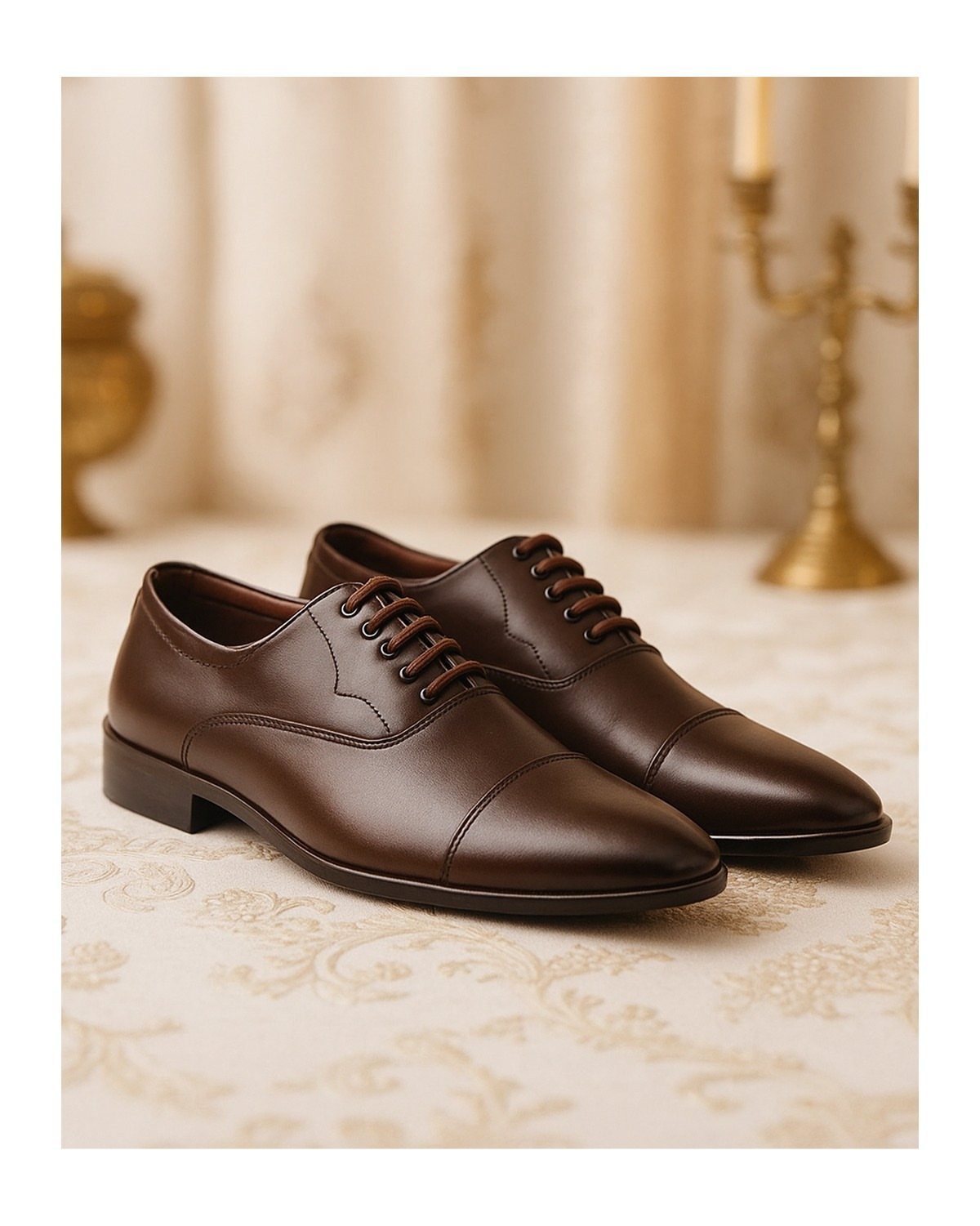 Brown leather men's dress shoes on a cream patterned tablecloth with gold accents.