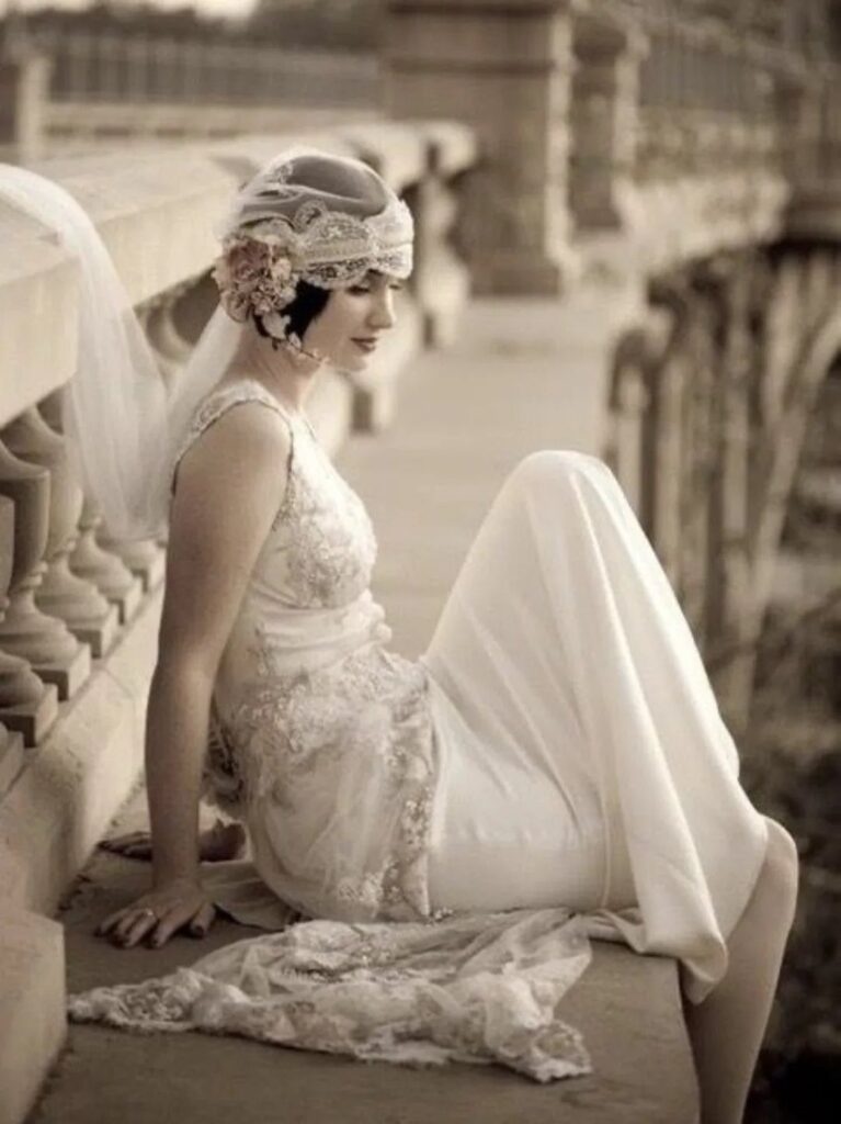 Woman in a beaded 1920s wedding dress and headpiece sitting on stone railing.