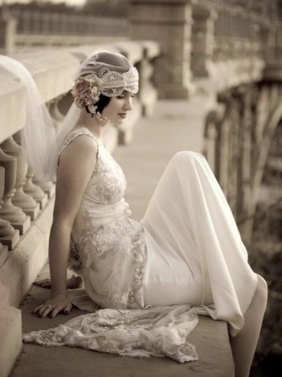Woman in a beaded 1920s wedding dress and headpiece sitting on stone railing.