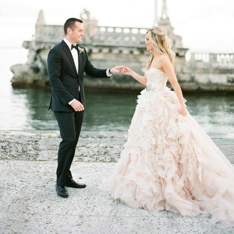 Bride in ruffled pink wedding dress and groom in black tuxedo holding hands.