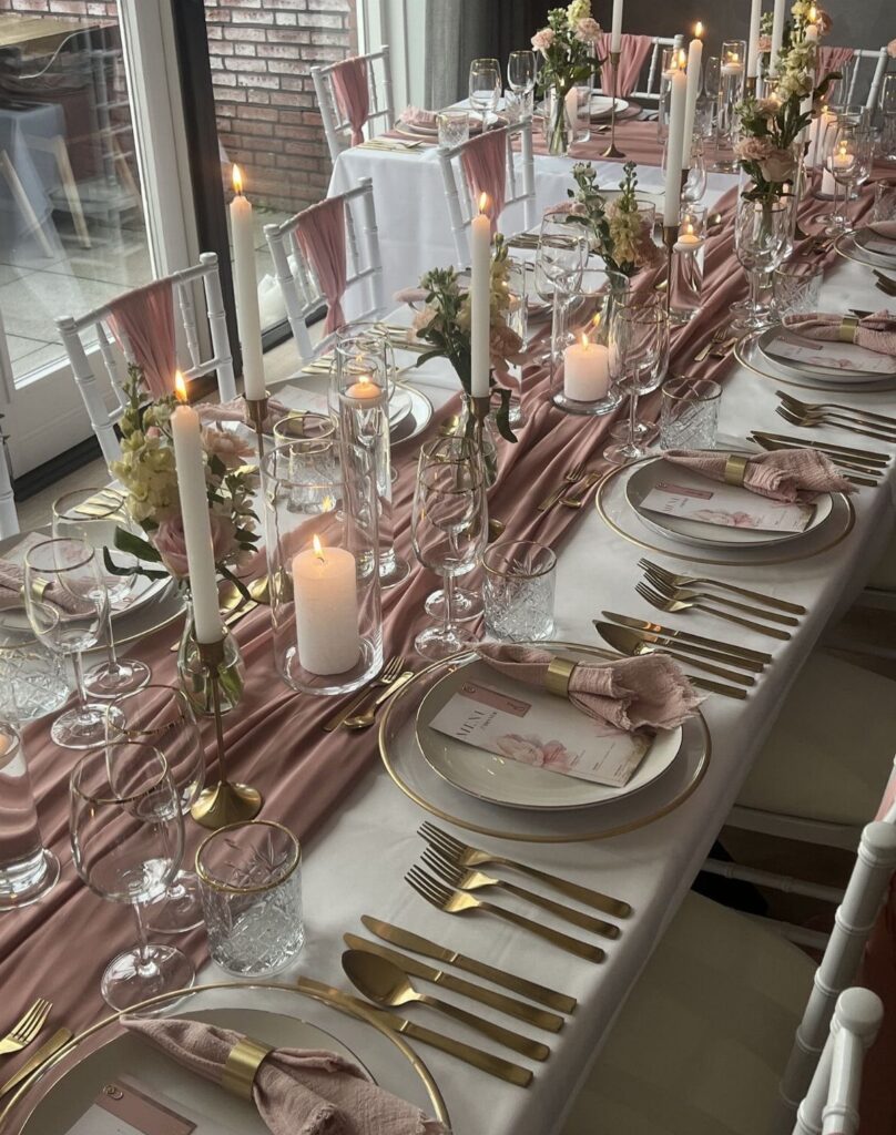 Wedding guest table with white linens, rose runner, gold cutlery, and white candles.