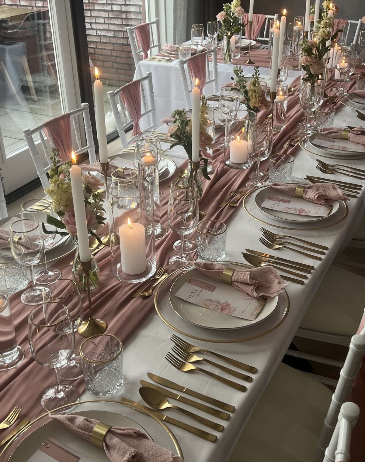 Wedding guest table with white linens, rose runner, gold cutlery, and white candles.