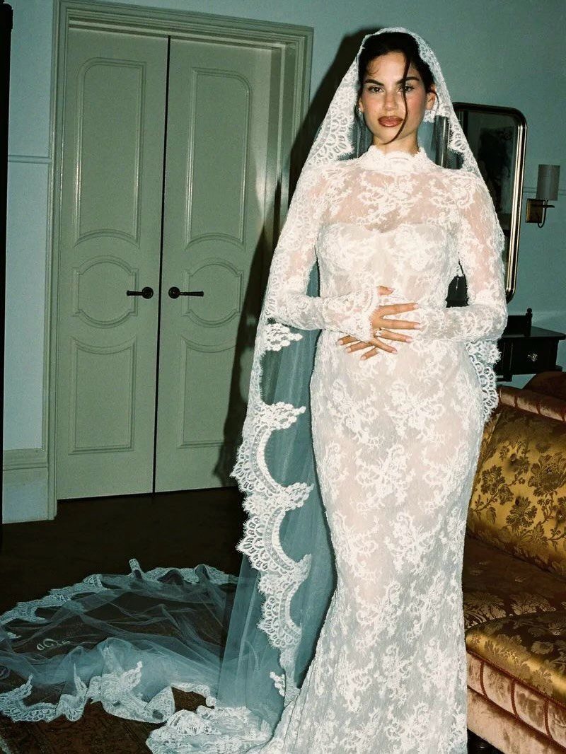 Woman in a long-sleeved white lace wedding dress and veil standing indoors.